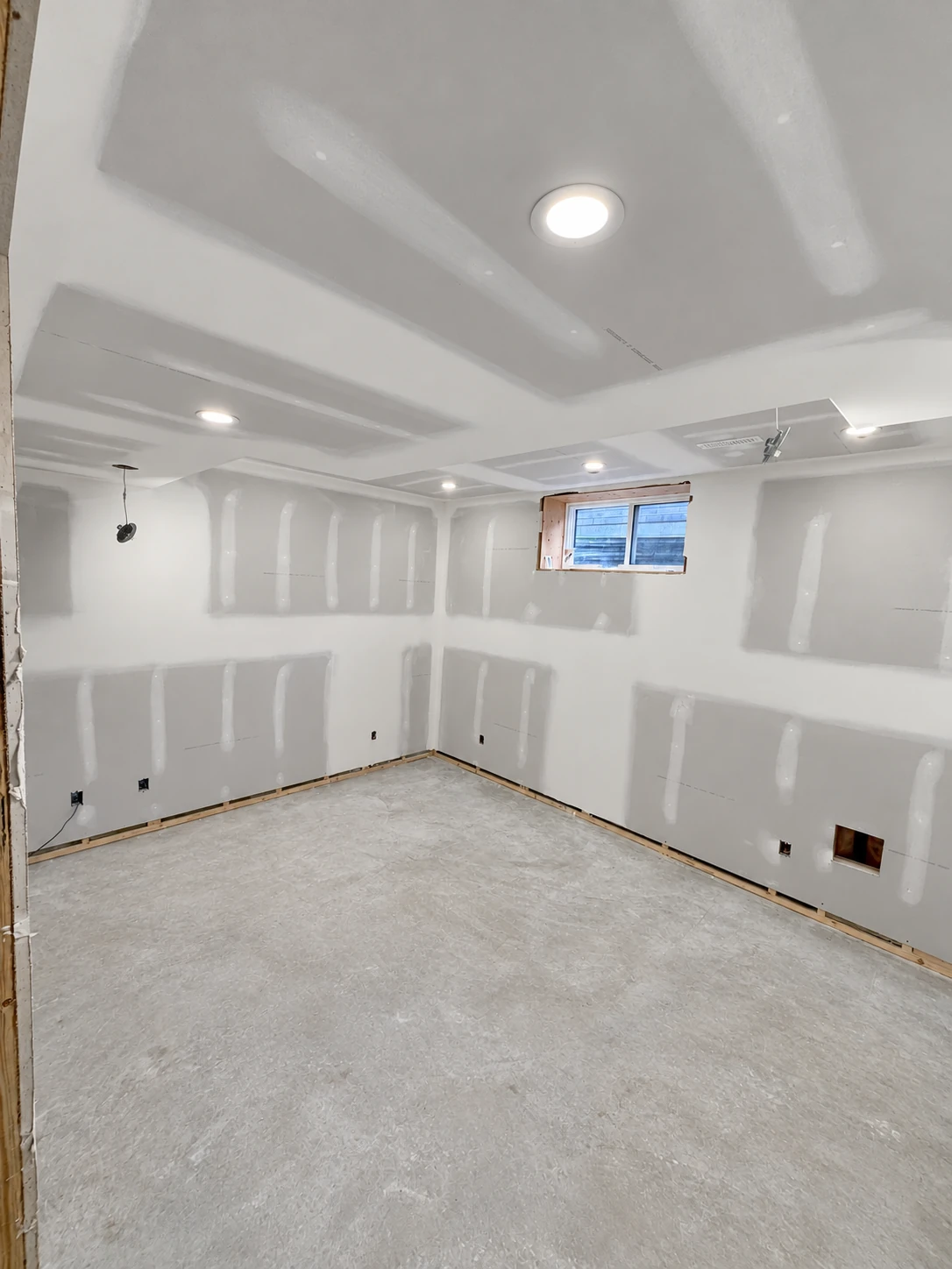 Unpainted basement drywall with broad compound bands across wall and ceiling joints beside a small window.